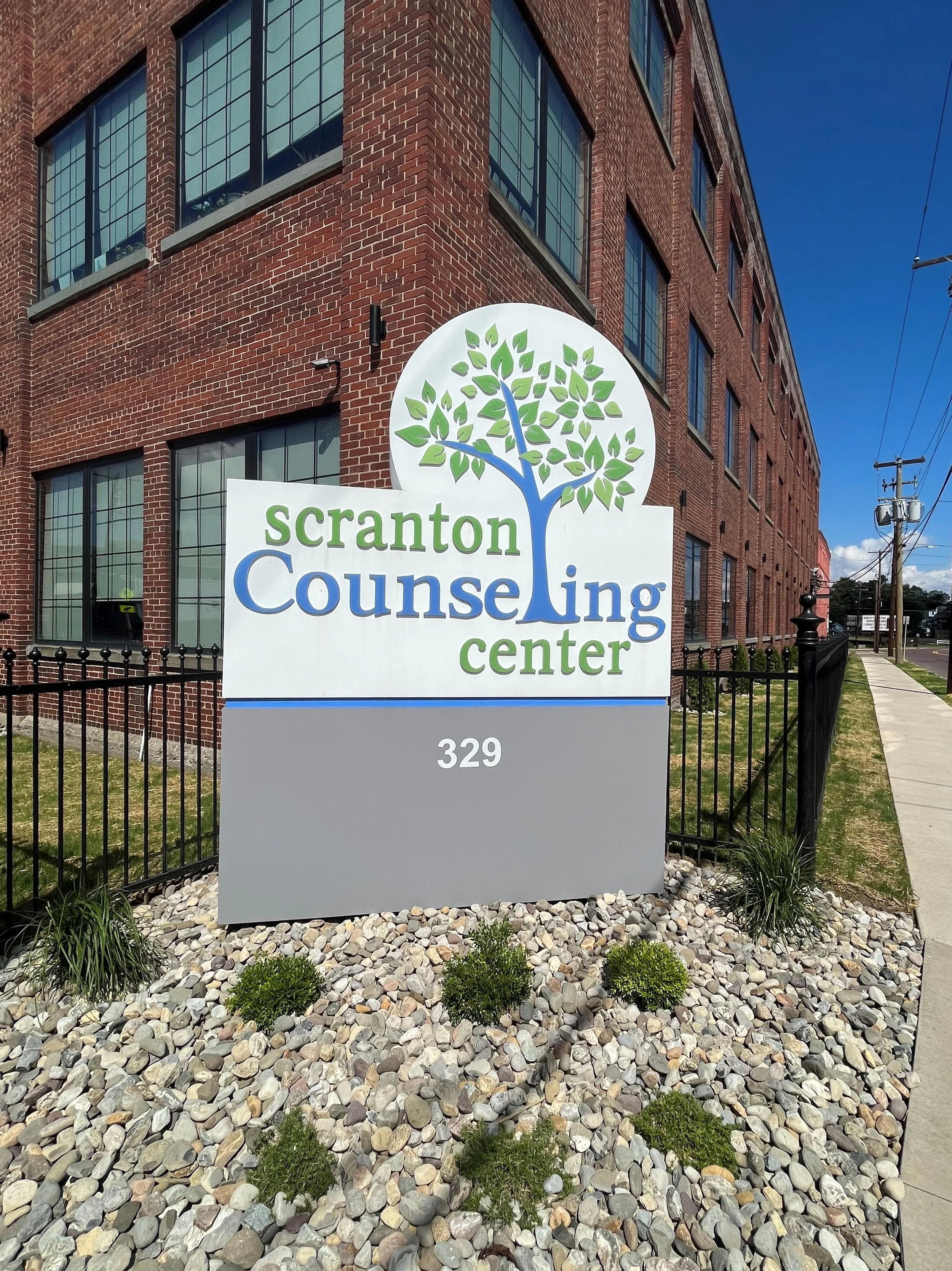 Sign for Scranton Counseling Center at 329, with a brick building in the background. The sign has a tree graphic with green leaves and blue branches, and the text in green and blue.