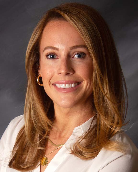 A woman with long, light brown hair wearing a white top and gold jewelry, smiling against a dark background.