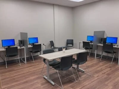 A room with four desks, each with a computer monitor and a chair, arranged against the walls, and a central table with chairs.