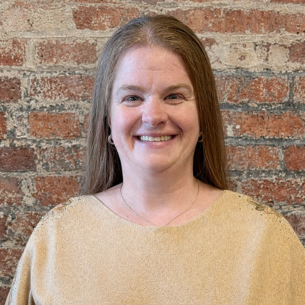 A woman with long brown hair, smiling, wearing a tan top against a brick background.
