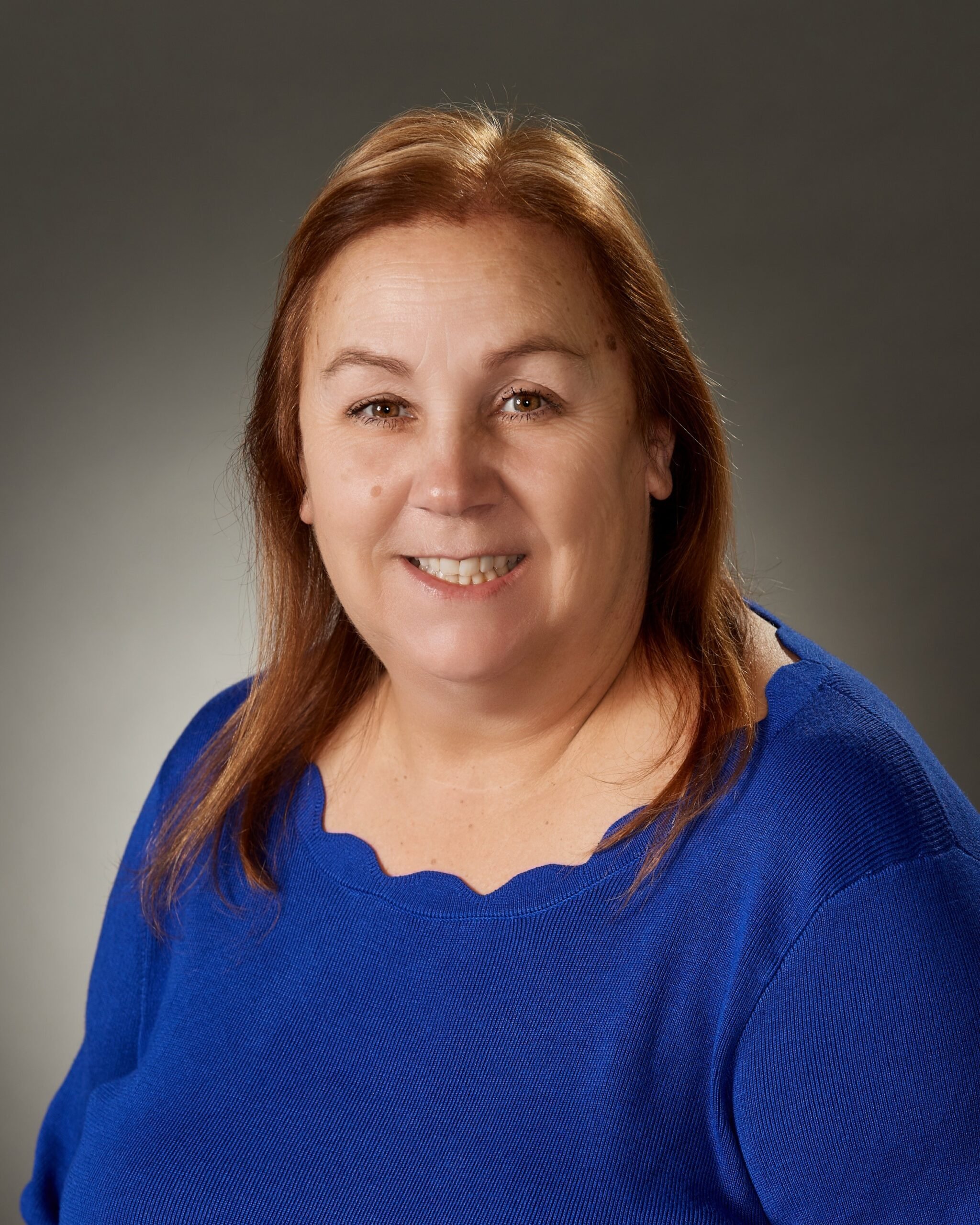A smiling middle-aged woman with shoulder-length red hair wearing a bright blue top against a dark gray background.