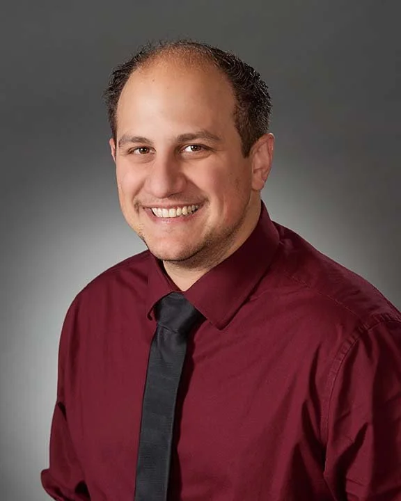 Headshot of a smiling man with short brown hair, wearing a maroon shirt and black tie, against a gray background.