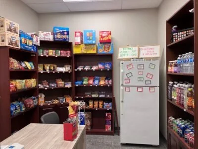 Small snack aisle with wooden shelves stocked with various snack boxes, a whiteboard with notes, and a table with snacks on it, in a room with plain walls.