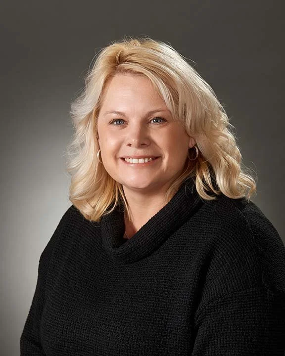 A smiling blonde woman with shoulder-length hair, wearing a black sweater, posing against a gray background.