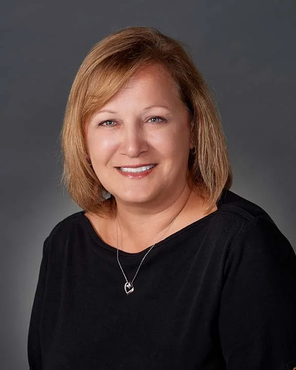 A woman with shoulder-length blonde hair, wearing a black top and a heart-shaped necklace, smiling against a gray background.