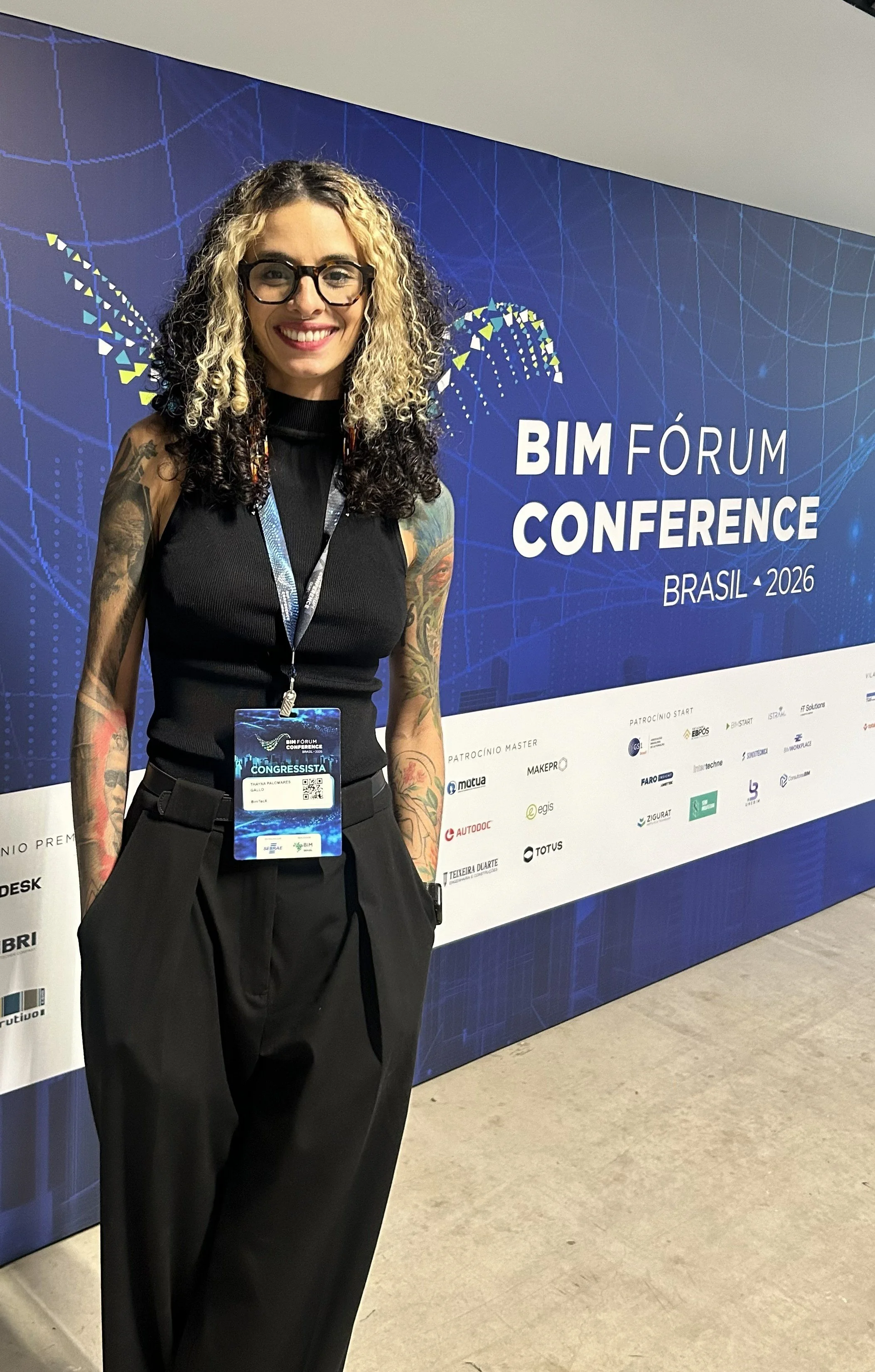 Mulher sorridente com cabelos cacheados, óculos de grau, tatuagens nos braços, usando roupa preta, com crachá, na conferência BIM Fórum Brasil 2026.