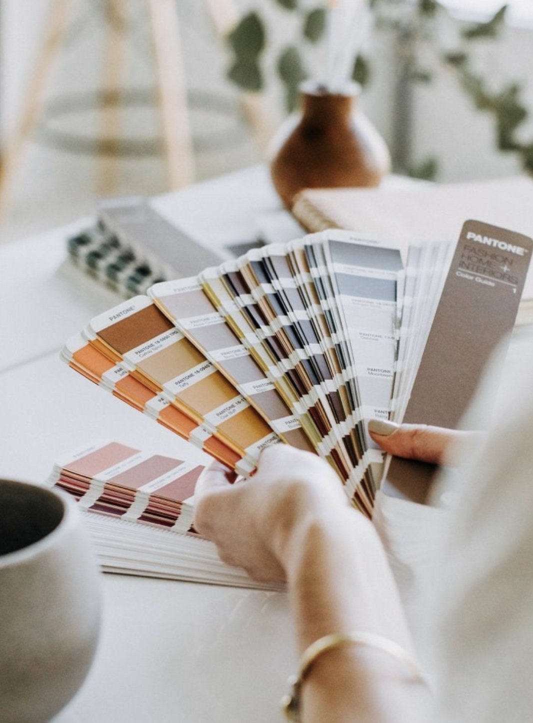 Mains tenant une palette de couleurs Pantone à fards diffrents, posée sur une table. En arrière-plan, une vase en céramique, un carnet, et un ordinateur portable.