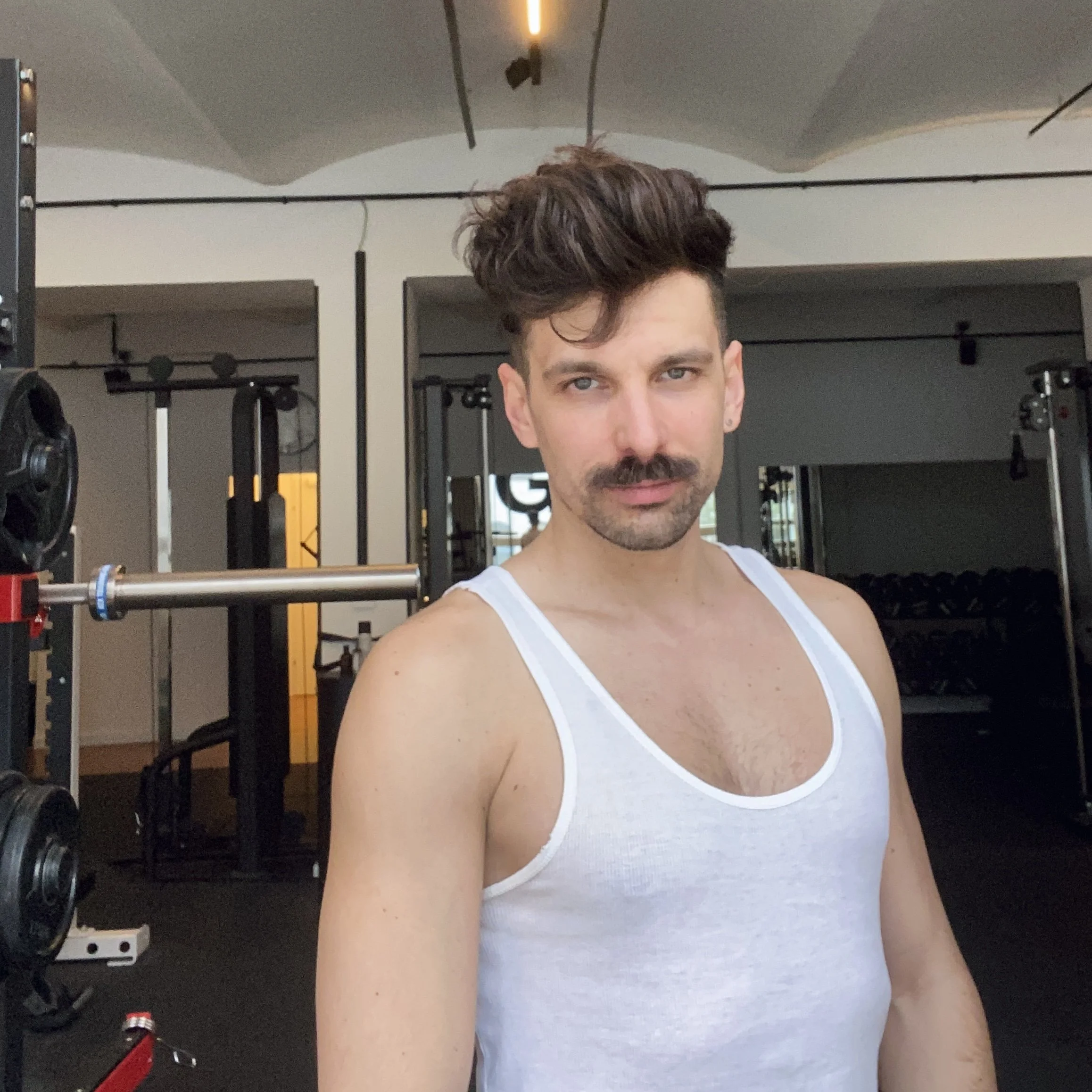 Mann mit weißem Tanktop und Schnauzer steht in einem Fitnessstudio und blickt direkt in die Kamera.