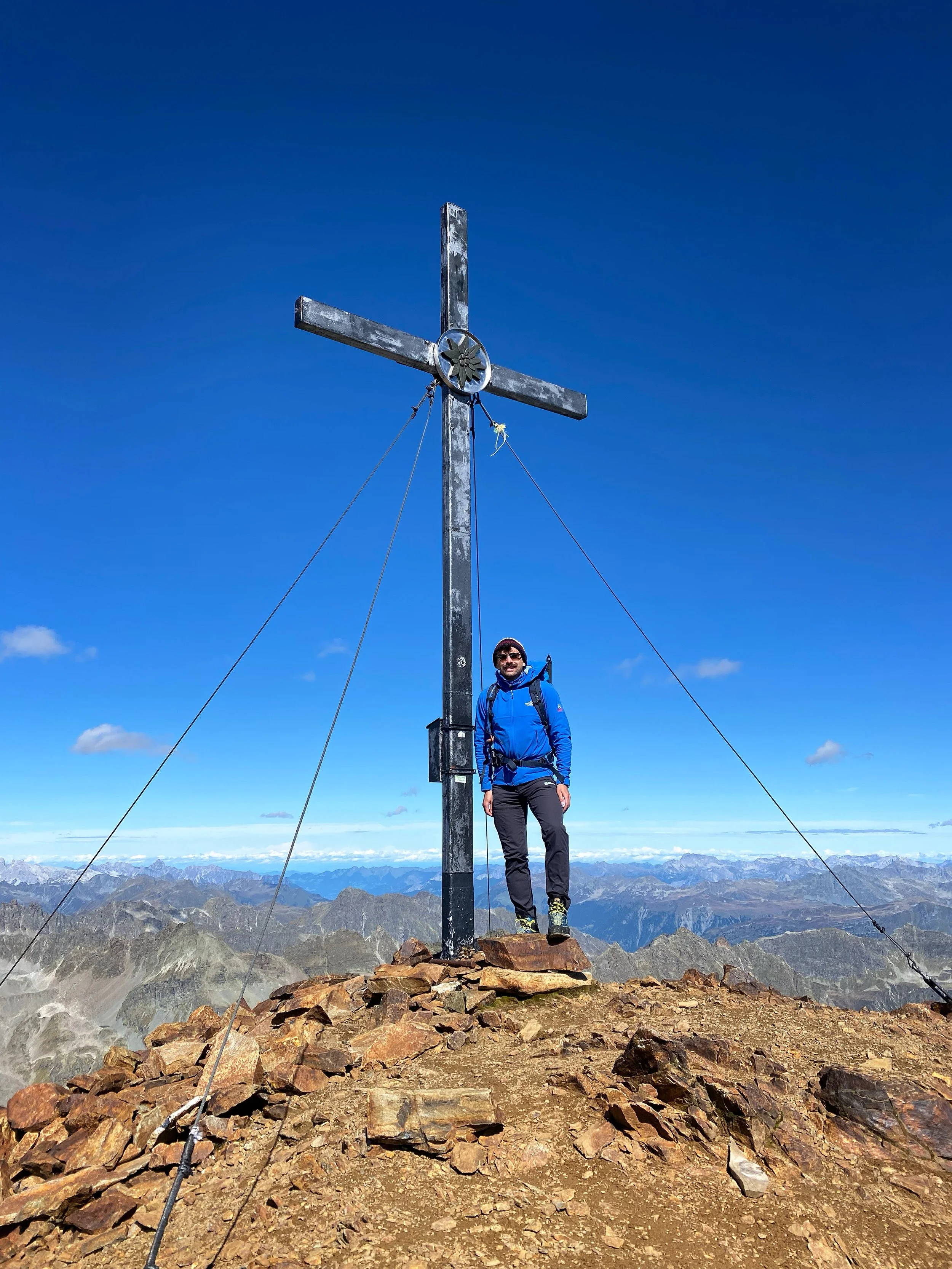 Mann in blauer Jacke steht auf dem Gipfel eines Berges vor dem Gipfelkreuz