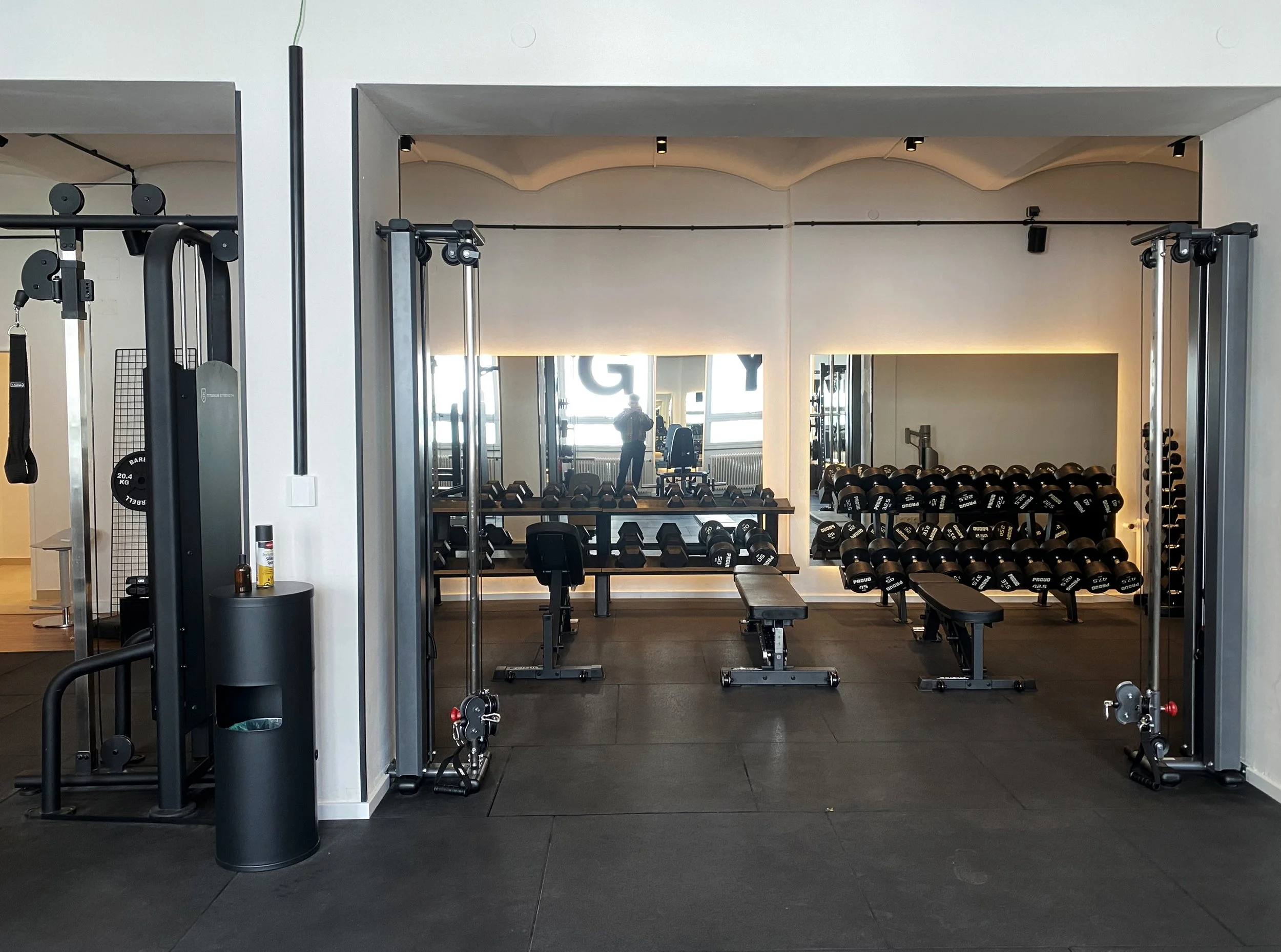 Innenaufnahme eines Fitnessstudios mit Hantelbänken, Kabelzügen und Kurzhanteln auf Regalen