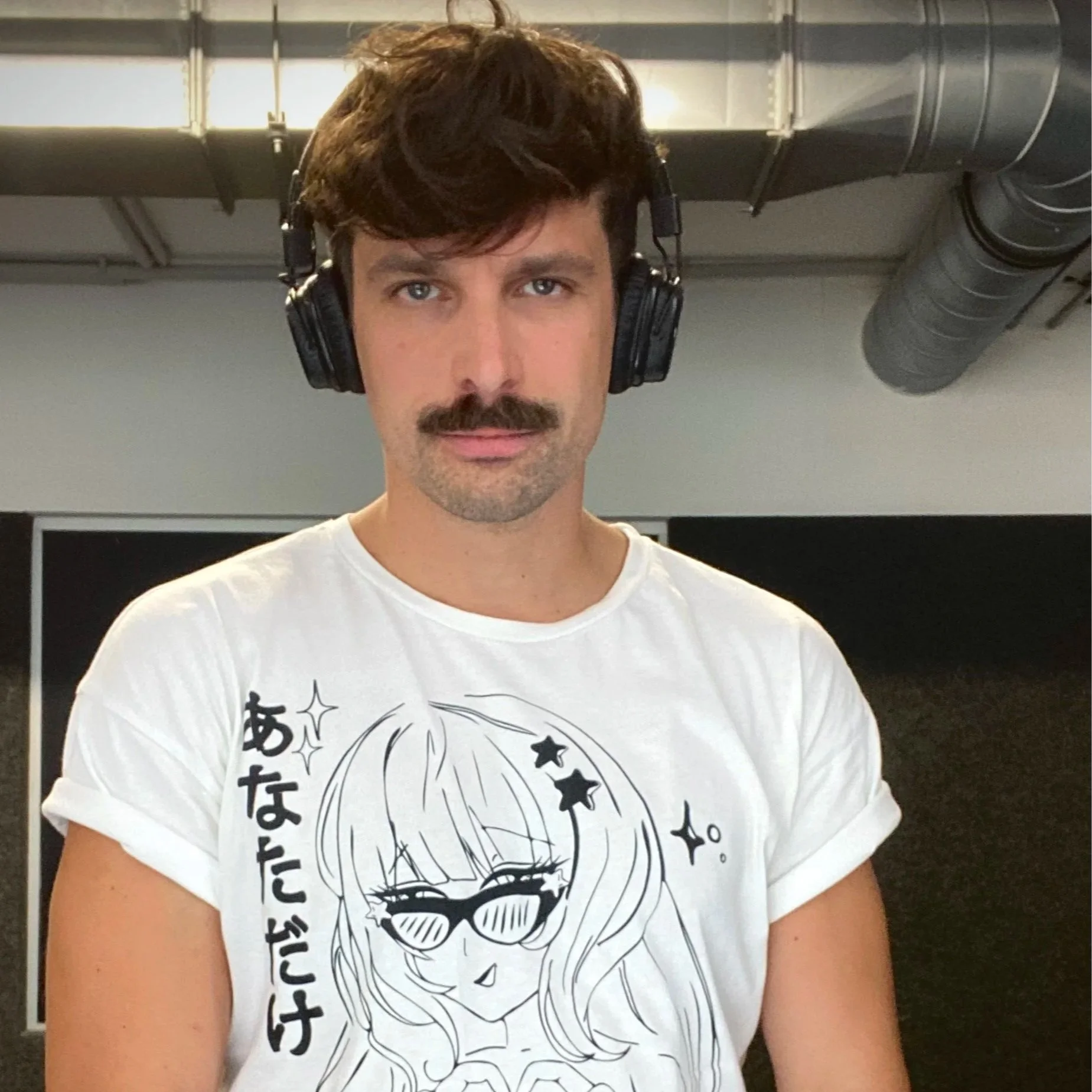 Junge mit dunklem Haar und Schnurrbart trägt Kopfhörer und ein weißes T-Shirt mit einem Manga-Charakter und japanischem Text, im Hintergrund industrielle Lüftungsschäuche in einem Raum.