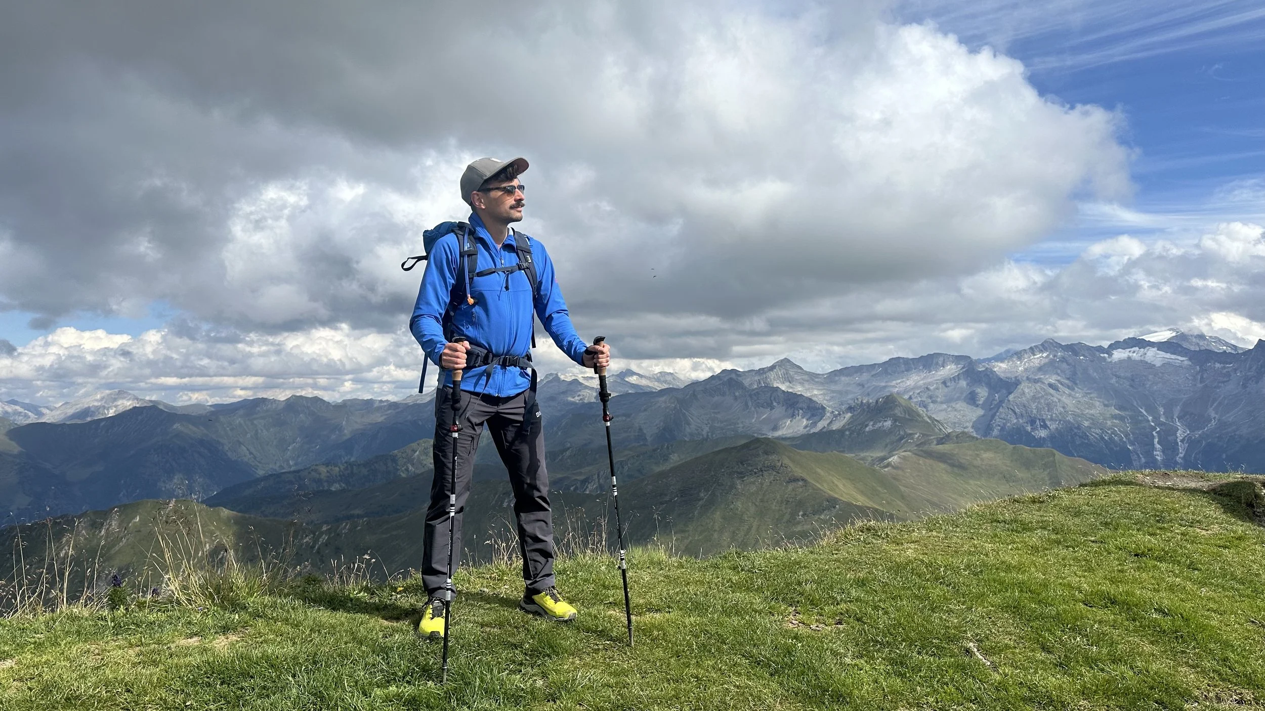 Mann trägt eine blaue Jacke, Kappe und Sonnenbrille und steht auf dem Gipfel eines Berges