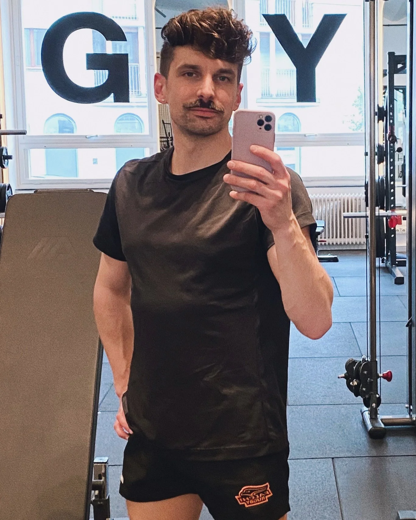 Mann mit leicht gelocktem Haar und Schnauzer steht vor dem Spiegel in einem Fitnessstudio und macht ein Selfie mit dem Smartphone