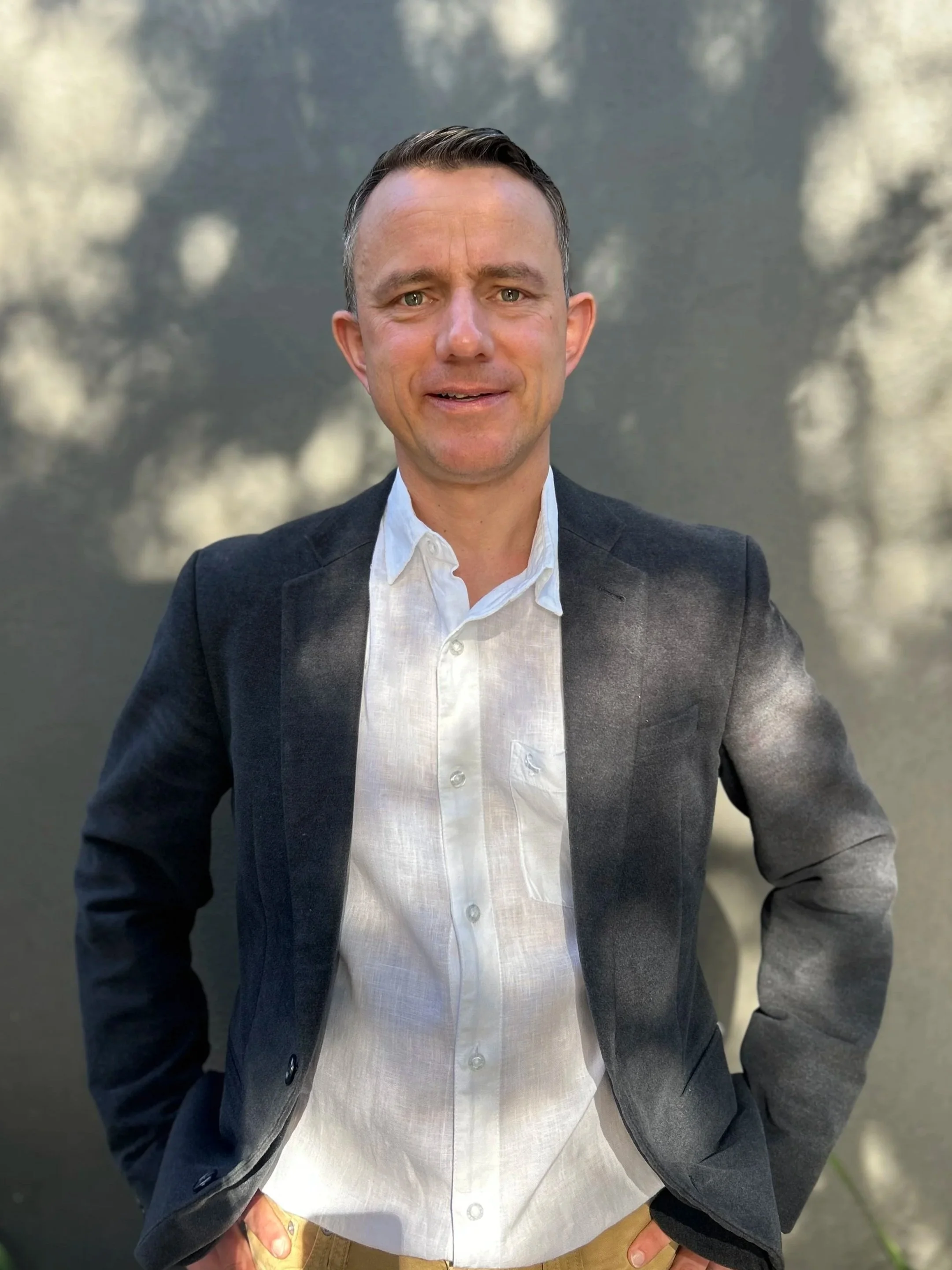 A man wearing a dark blazer, white shirt, and khaki pants standing outdoors with a blurred natural background.