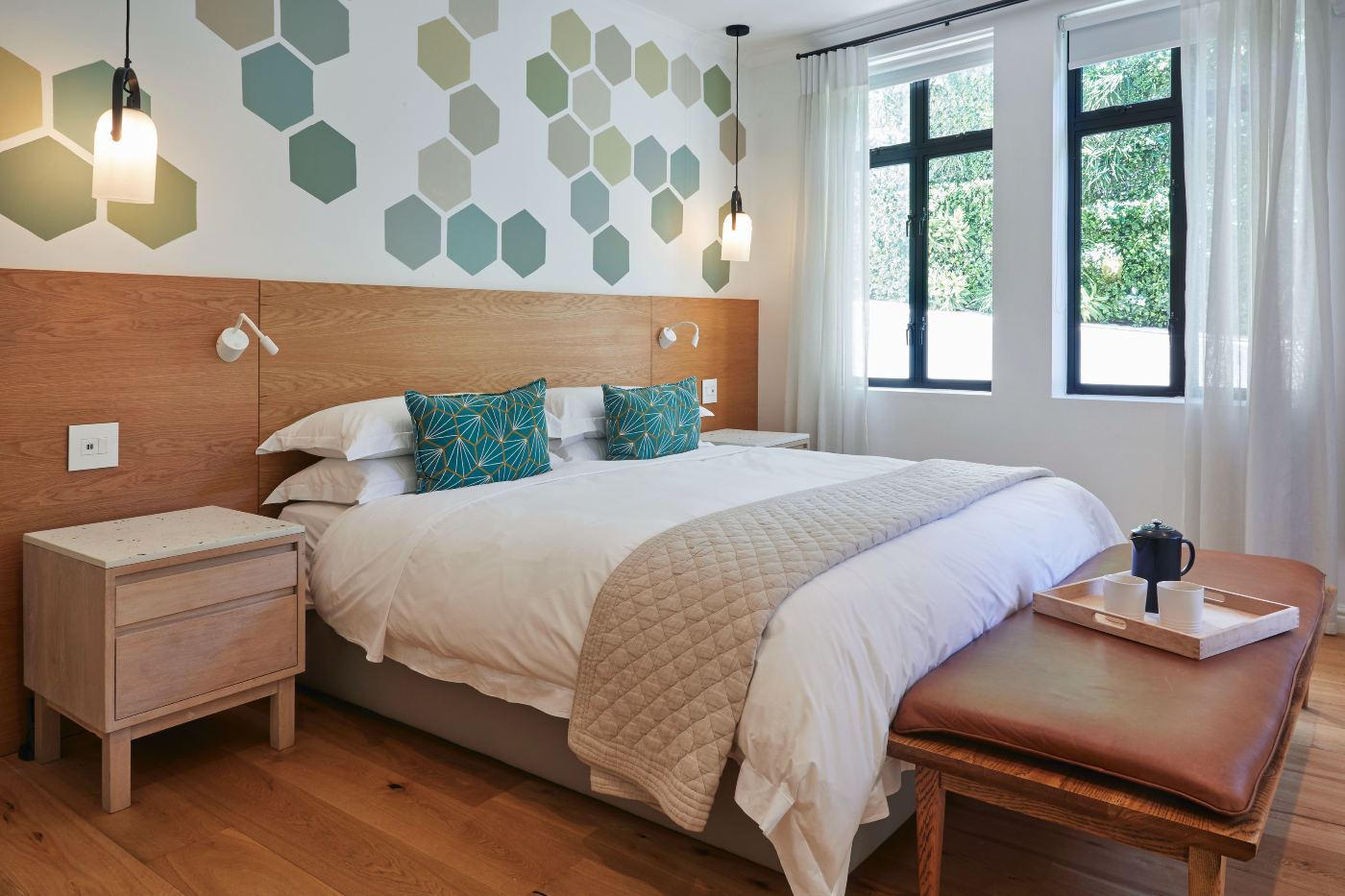 A cozy bedroom with a large bed featuring white bedding, two blue patterned pillows, and a beige quilt. There are two hanging pendant lights above the bed, a wooden headboard, and two wall-mounted reading lights. Natural light streams through three big windows with black frames and sheer white curtains. Next to the bed are white nightstands, one with a small drawer. In front of the bed, there is a wooden bench with a leather cushion, holding a tray with a teapot and cups.