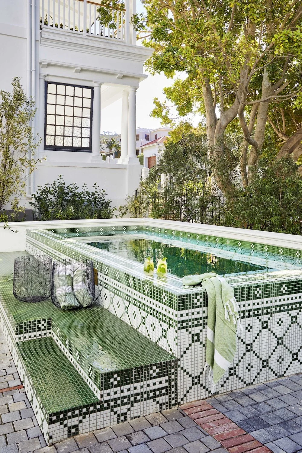 A backyard with a mosaic-tiled hot tub, two green drinks, and a towel, surrounded by greenery and residential buildings.