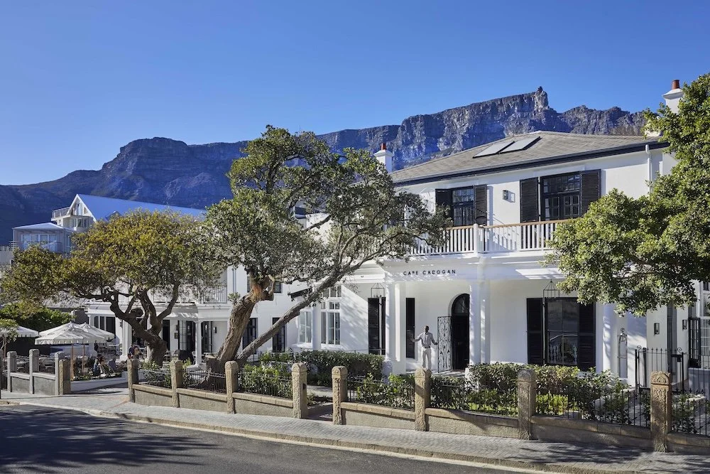 White building with black shutters and a balcony, surrounded by trees, with mountains in the background.