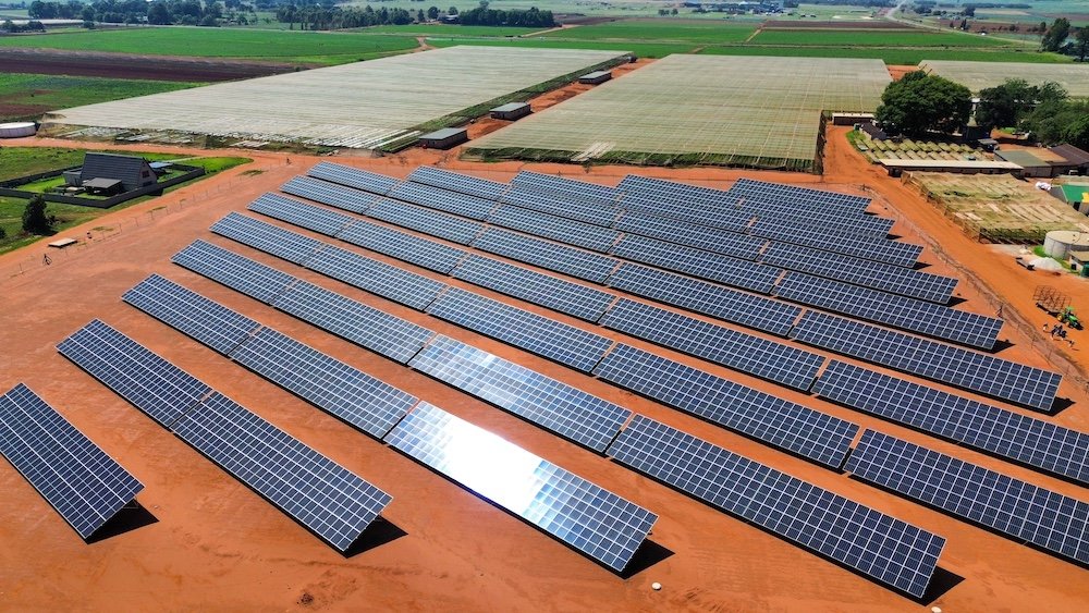 Several rows of solar panels installed on red dirt land in a rural area with farmland and green fields surrounding the site.