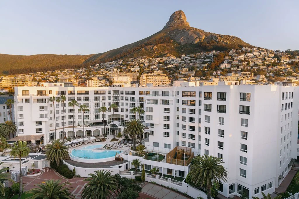 White hotel building with a swimming pool surrounded by palm trees in a hilly city landscape with a mountain in the background.
