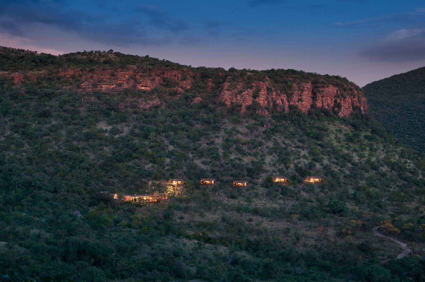 Lights on houses or cabins situated on a hillside in a mountainous landscape during dusk, with a large rocky formation at the top and a cloudy sky above.
