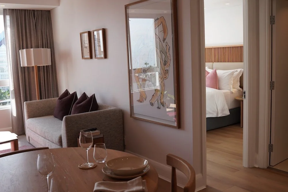 Interior of a modern hotel room with a sitting area and a view into a bedroom; decorated with framed art, tableware, wine glasses, pillows, a bed, and wooden accents.