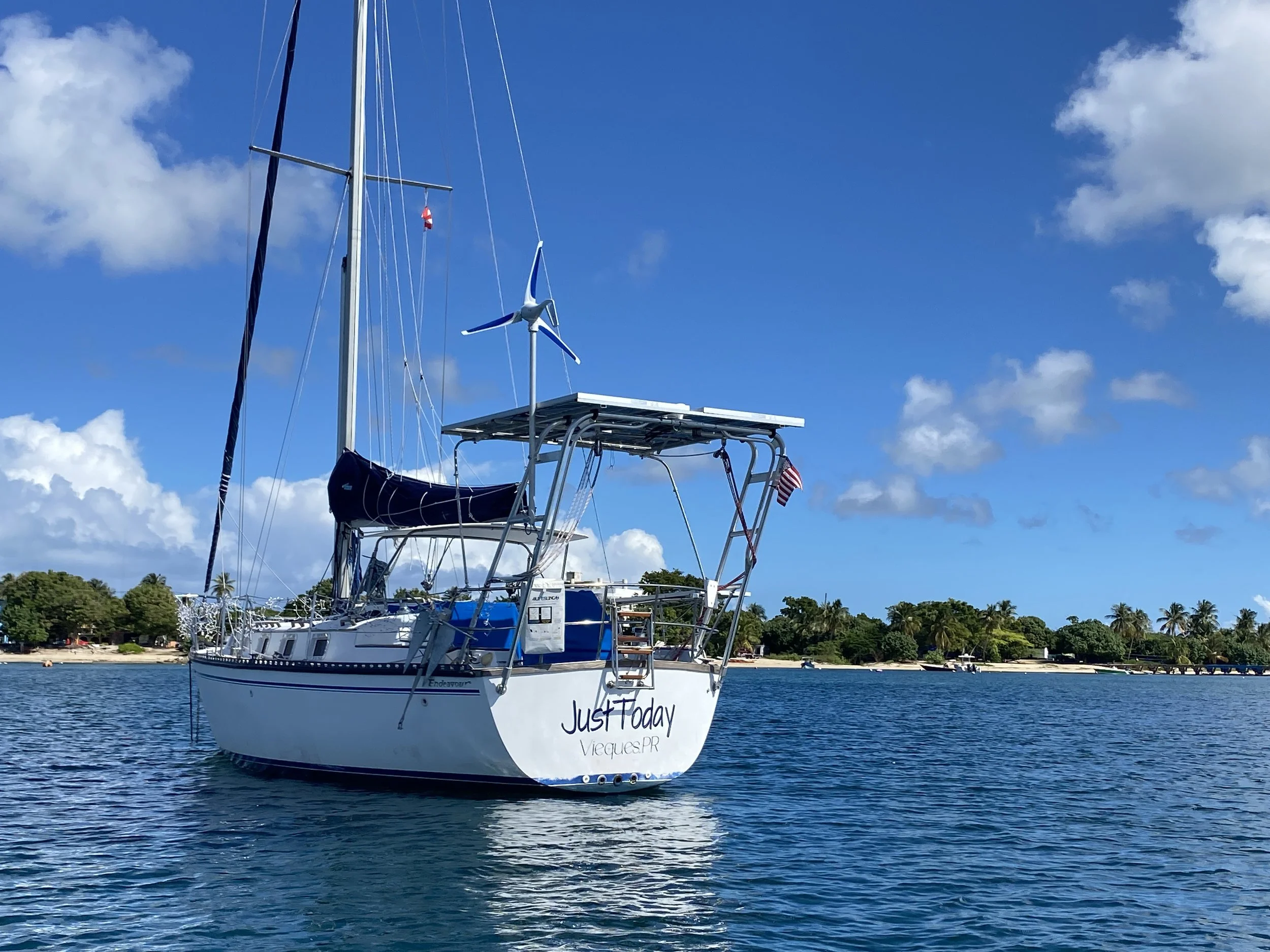 A sailboat named 'Just Today' on calm water near a shoreline with trees and palm trees under a partly cloudy sky. The electric drive boat has a solar panel and a wind turbine on top.