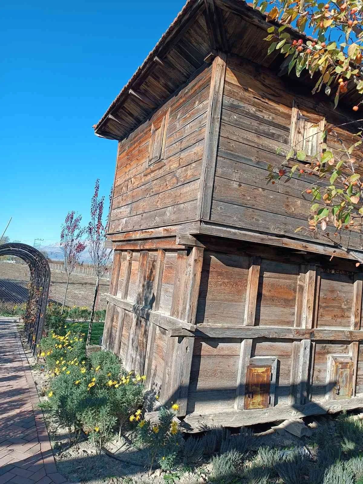 Ahşap eski bir köy evi veya kulübe, dış bahçede çiçekler ve yürüyüş yolu, gökyüzü berrak ve mavi.