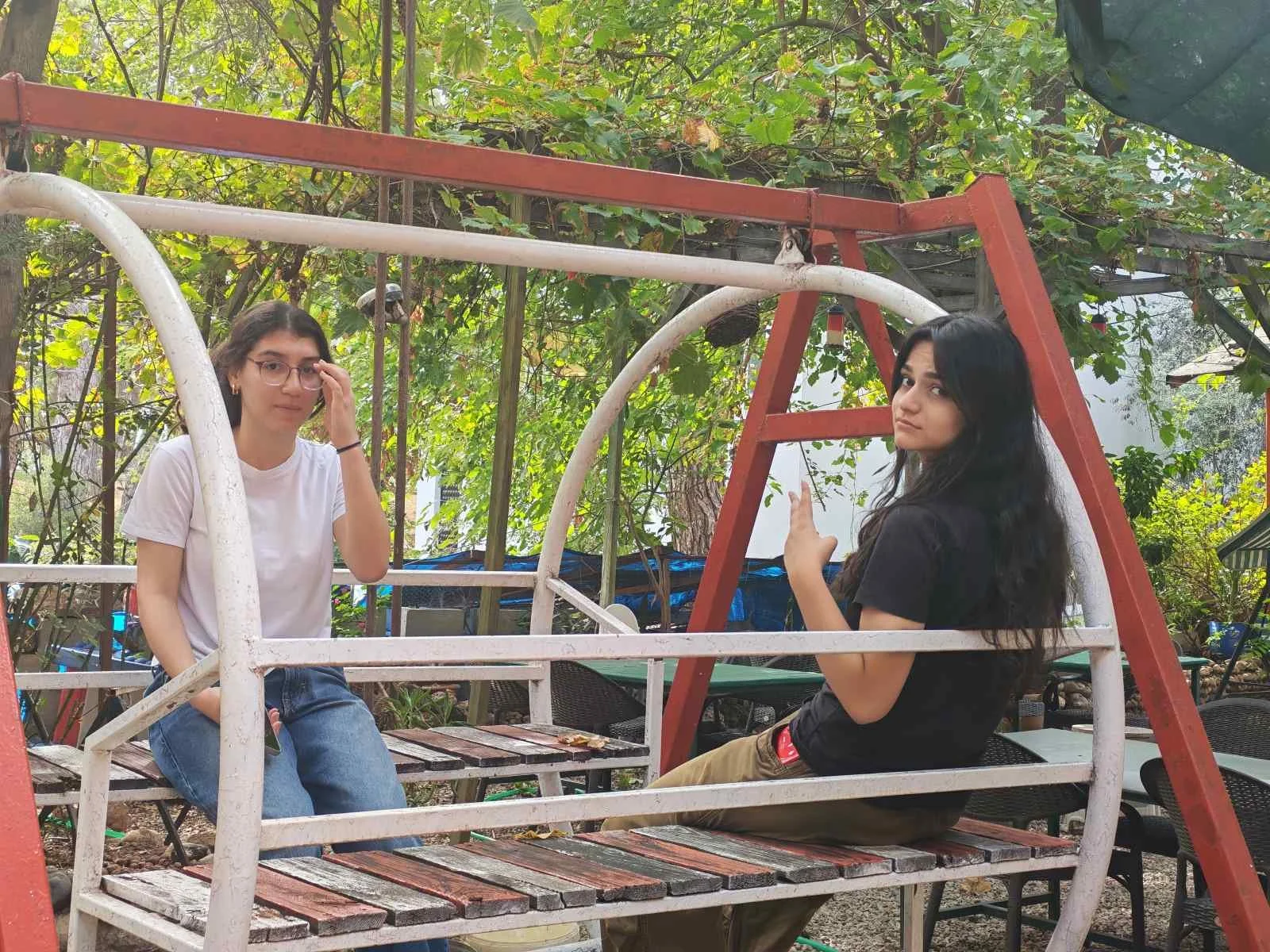 İki genç kadın, parkta kızaklı bir salıncağın üzerinde oturuyor, arka planda ağaçlar ve bitki örtüsü var.