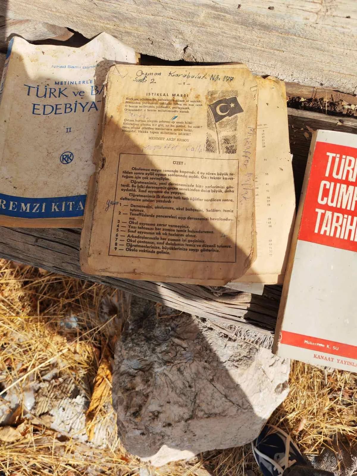 Eski ve yıpranmış kitaplar açık bırakılmış, üzerinde Türk milli marşı ve eğitimle ilgili yazılar var. Kitaplar ve taşın yer aldığı açık alanda kuru otlar ve taşlar bulunuyor.