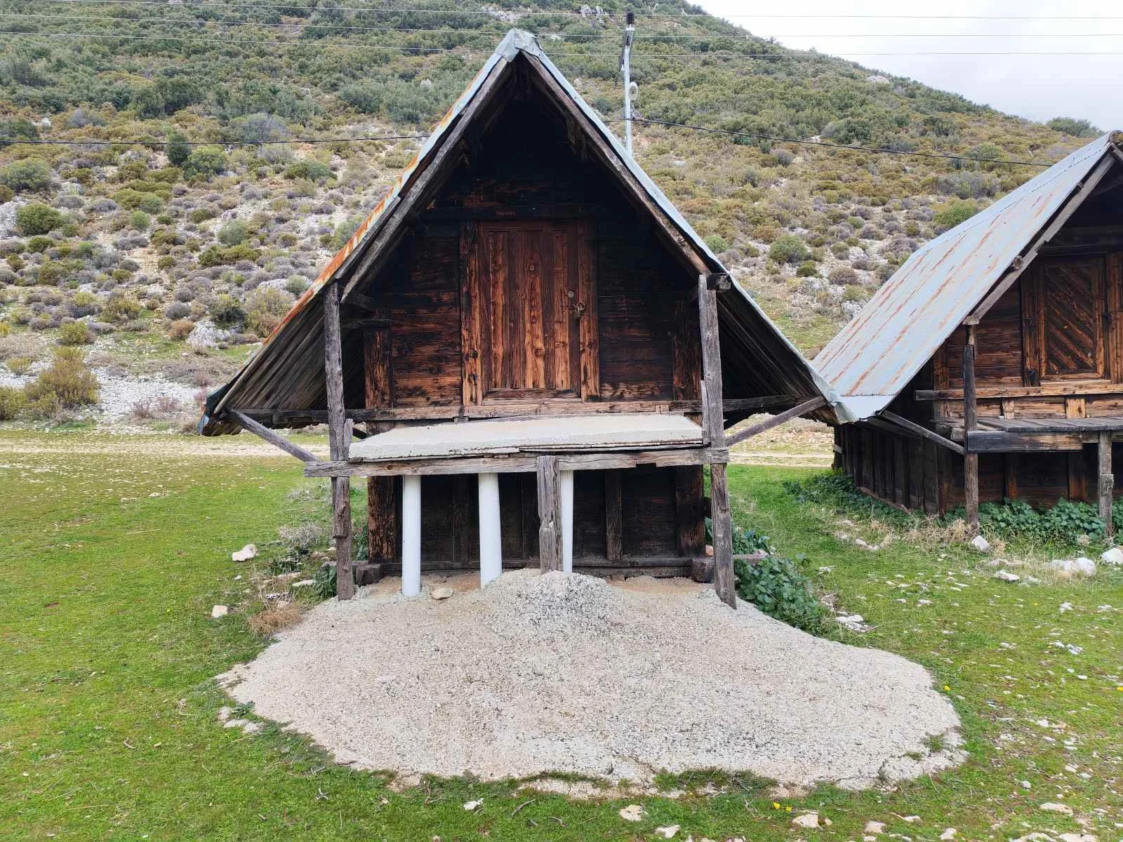 Dağlık alanda ahşap evler. Birisi yükseltilmiş ve çatısı metal, diğerki ise klasik ahşap yapı. Çimenlik ve taşlar ortada, arka planda ise dağlar ve çalılık bulunuyor.