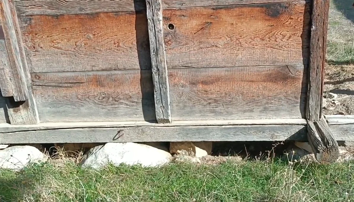 Eskimiş ahşap bir yapı ve yapının altındaki büyük taşlar ile çimlerin görünümü.