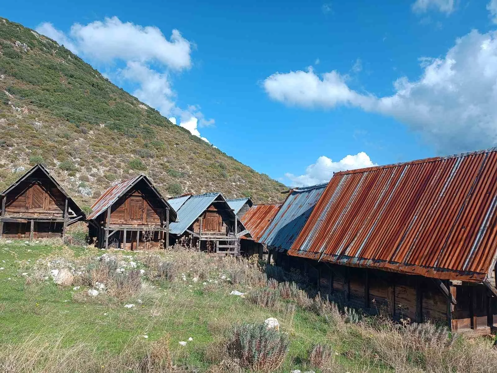 Dağlık alanda, parça parça rustik ahşap evler ve çatılarıyla küçük köy. Hava açık ve gökyüzü mavi, bazı beyaz bulutlar var.