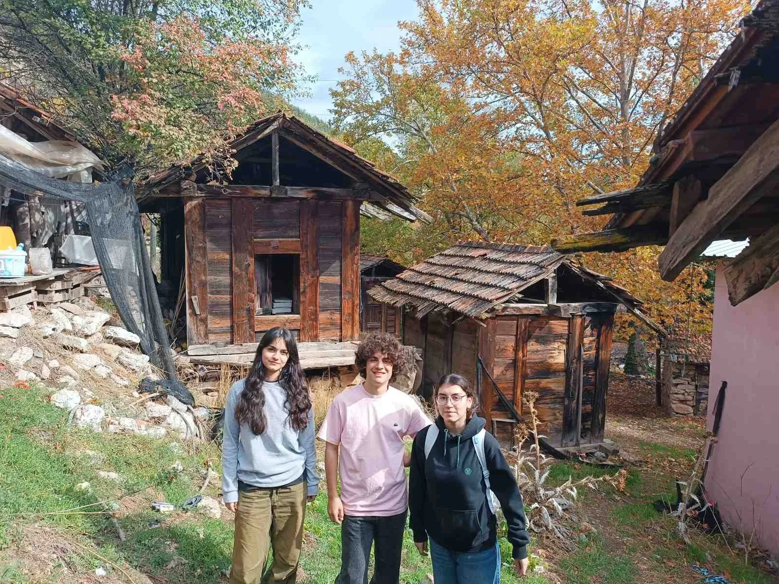 Açık hava köyünde üç kişi, arka planda eski ahşap yapılar ve renkli sonbahar ağaçları ile güzel bir doğa manzarası.