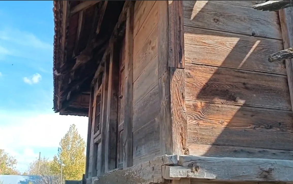 Ahşap eski bir evin köşesi, ahşap duvarlar ve çatının detayları. Arka planda gökyüzü ve ağaçlar visible.