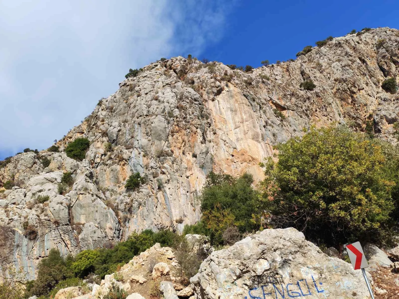 Sırtında büyük kaya ve bazı ağaçlar bulunan dağ manzarası, önünde yazılmış 'Şanslı' kelimesi bulunan taş ve sağda kırmızı beyaz dönüş levhası.