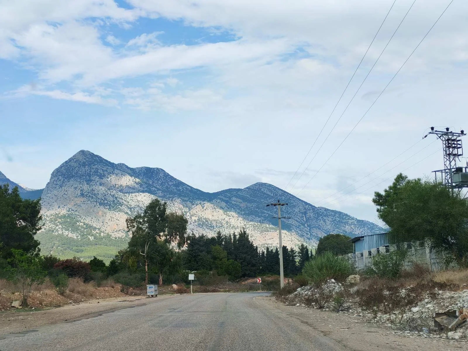 Dağlar ve bulutlu gökyüzü ile bir kırsal yol, yol kenarında elektrik direkleri ve bazı ağaçlar var.