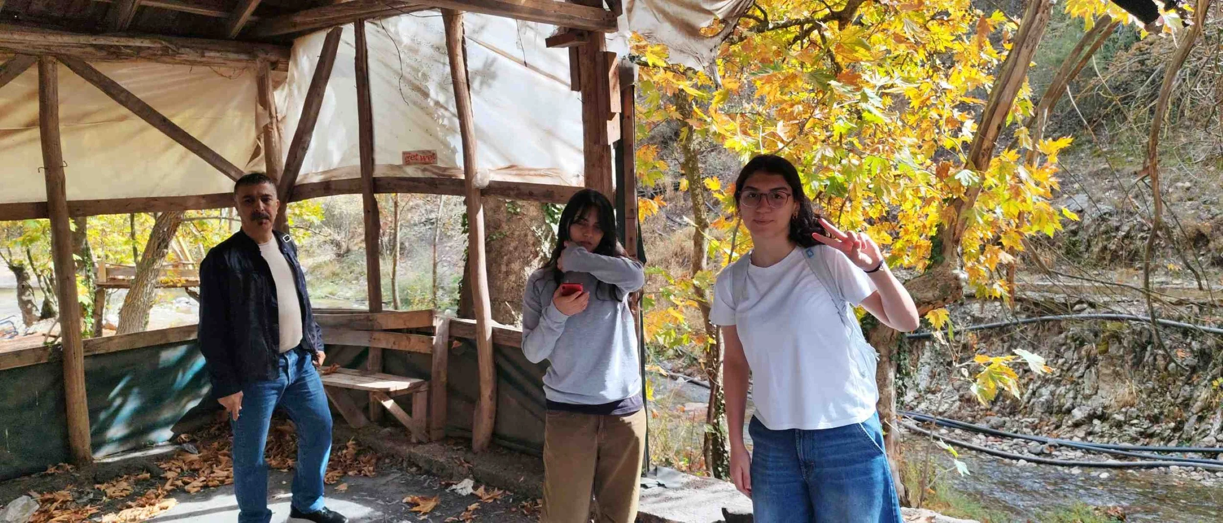 Ağaçlık alanda, üç kişi bir arada. Sol tarafta, erkek ve kız. Sağda, kız. Arka planda ağaçlar ve yapraklar, sonbahar mevsimi, yapraklar sarı ve kahverengi.