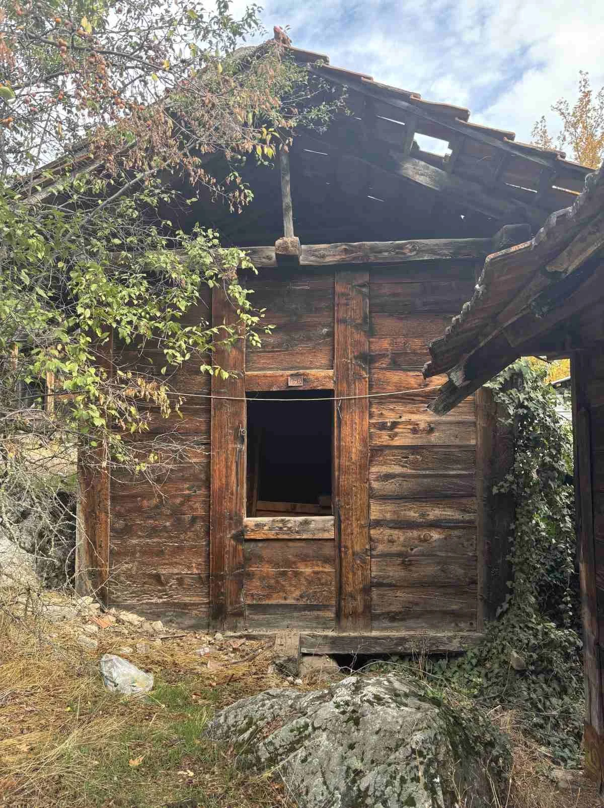 Eski ve bakımsız ahşap bir kulübe. Bir pencere ve çatısında hasar var. Kulübenin önünde taşlar ve bazı bitkiler var.