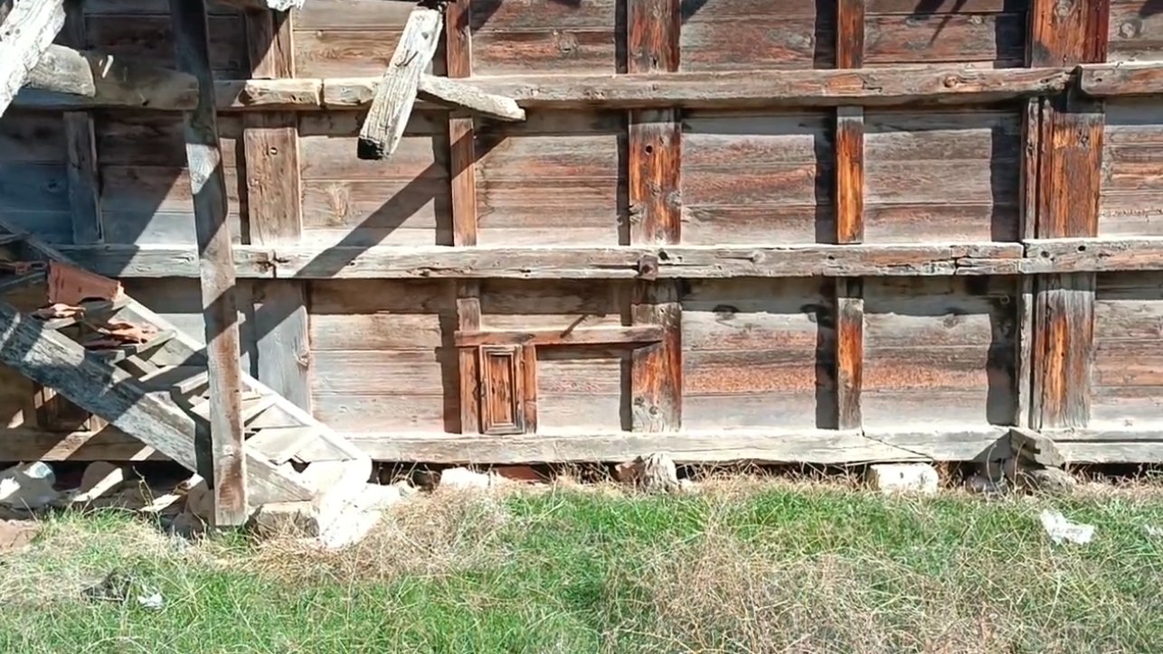 Ahşap bir duvar ve önünde toprak ve çim alan, duvarın sol köşesinde ahşap merdiven ve basit yapım araçları bulunuyor.