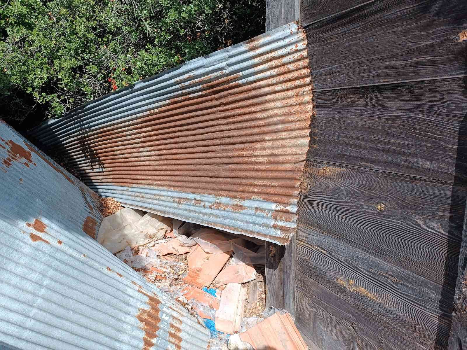 Eski ve paslı metal çit, ahşap zemin ve duvar ile çevrili, arkasında bitki bulunan boş ve atık dolu alan.