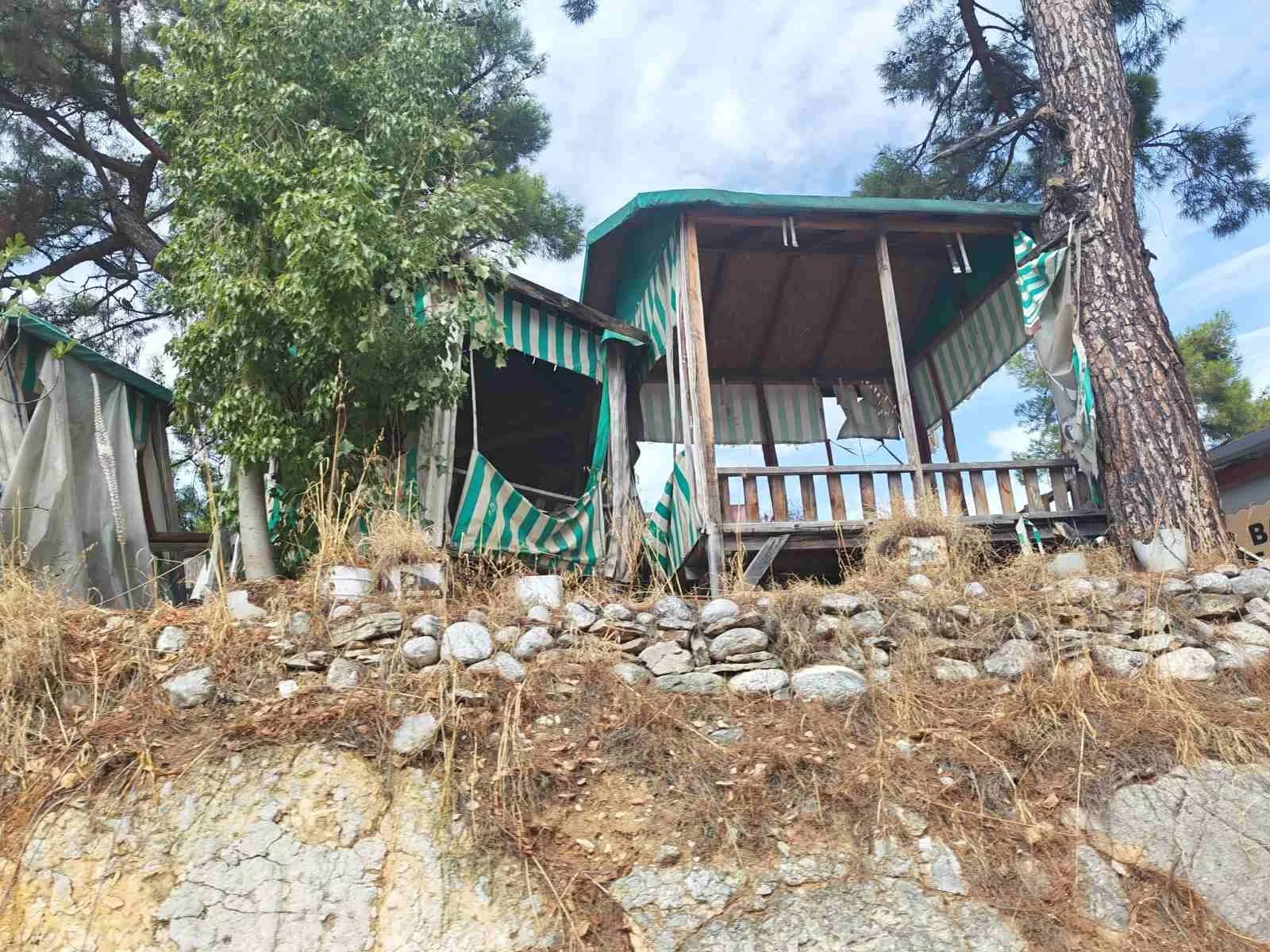 Yıkılmış ve bakımsız ahşap iskeletli çardak ve küçük kulübe, etrafında kurumuş otlar ve taş duvarlar, büyük bir ağaç ve birkaç ağaç yaprağı arasında, mavi gökyüzü ve bulutlar görünmektedir.