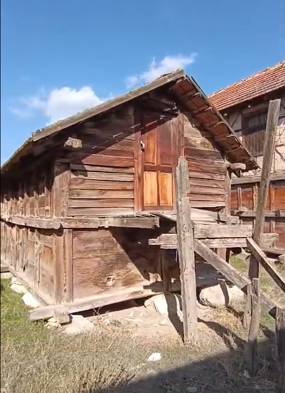 Ahşap geleneksel ev, ahşap merdivenle çıkış, mavi gökyüzü ve bulutlar, taş temel üzerinde duruyor.