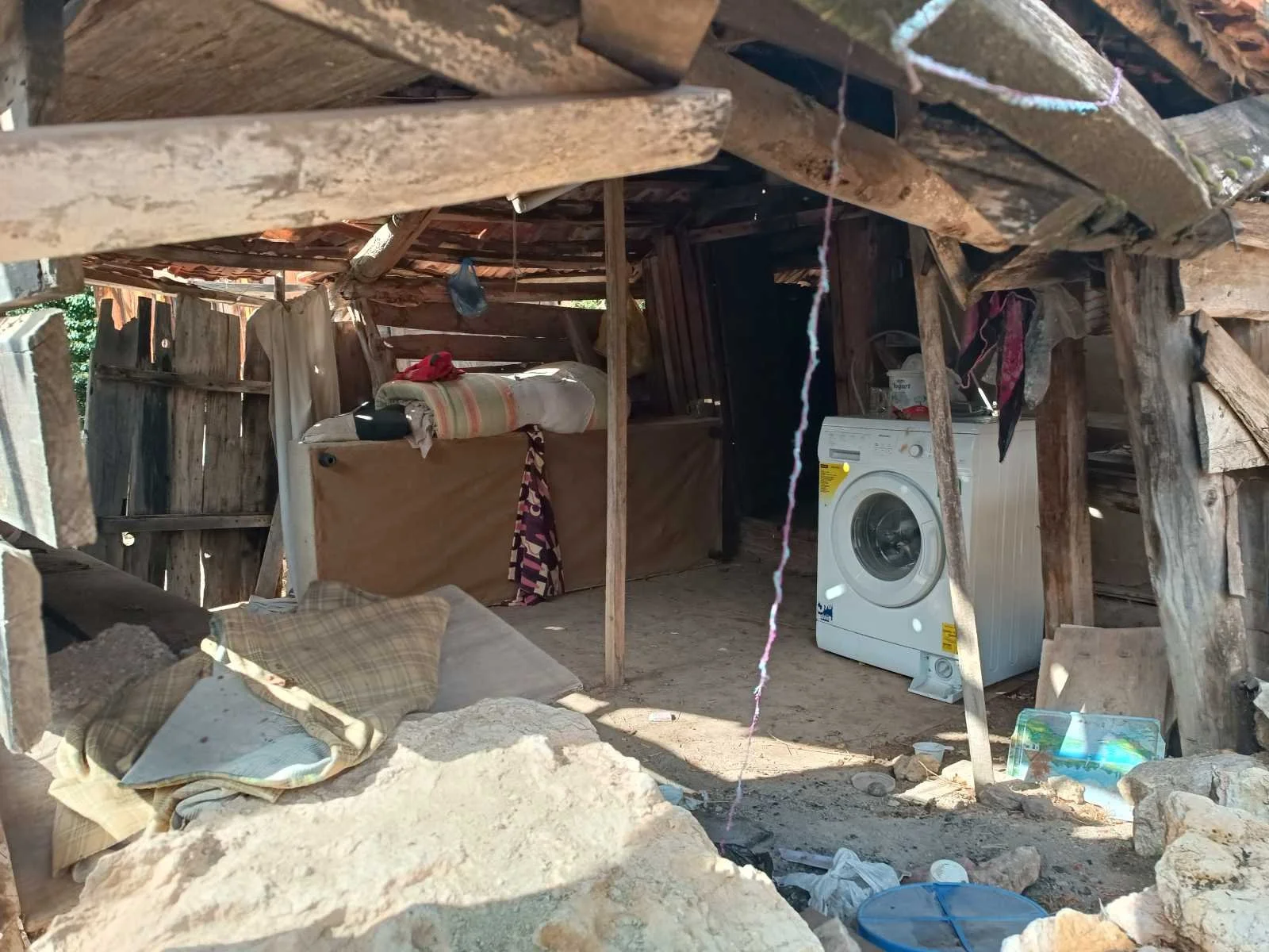 Dikenlikte, ahşap ve taşlardan yapılmış eski bir ev veya baraka, içerde bir çamaşır makinesi ve bazı eşyalar bulunmakta.