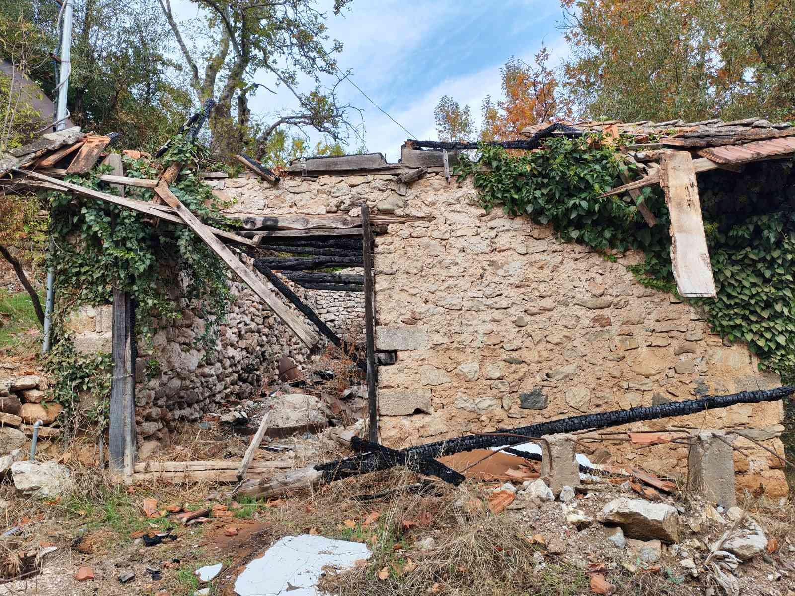 Yıkılmış bir taş ve ahşap duvar, hasar görmüş çatılar ve etrafta yayılmış molozlar ile doğal çevrede tahrip olmuş bir yapı.