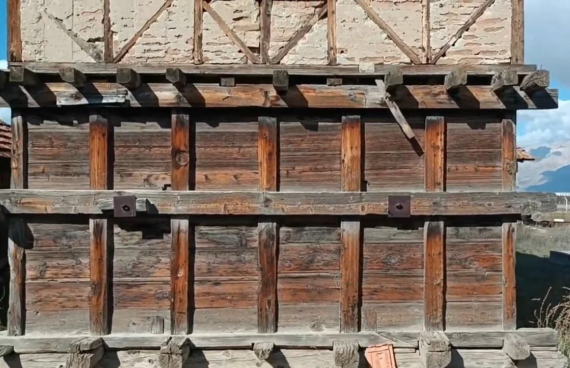 Ahşap yapı iskeleti, duvarlar ve destekler görünmekte, muhtemelen eski veya terkedilmiş bir bina. Üst kısımda betondan bir bölüm ve biraz mavi gökyüzü hakim.