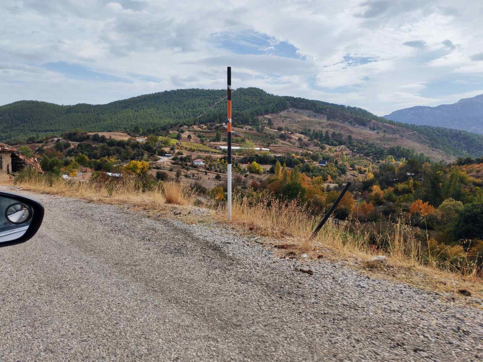 Dağlık alanda kırsal bir yolda ilerleyen araba, yol kenarında eğilmiş bir direk ve uzaklardaki yeşil tepeler ve ağaçlar.