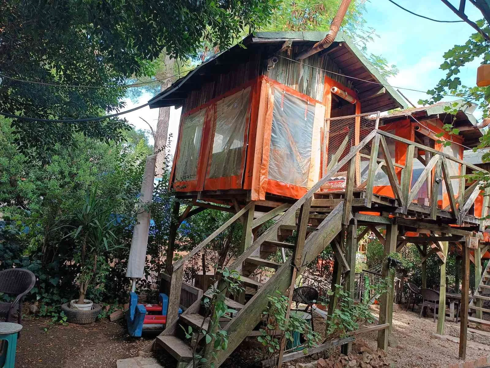 İki katlı ağaç ev, ahşap merdivenle ulaşılabilir, çevresinde ağaçlar ve açık alan.