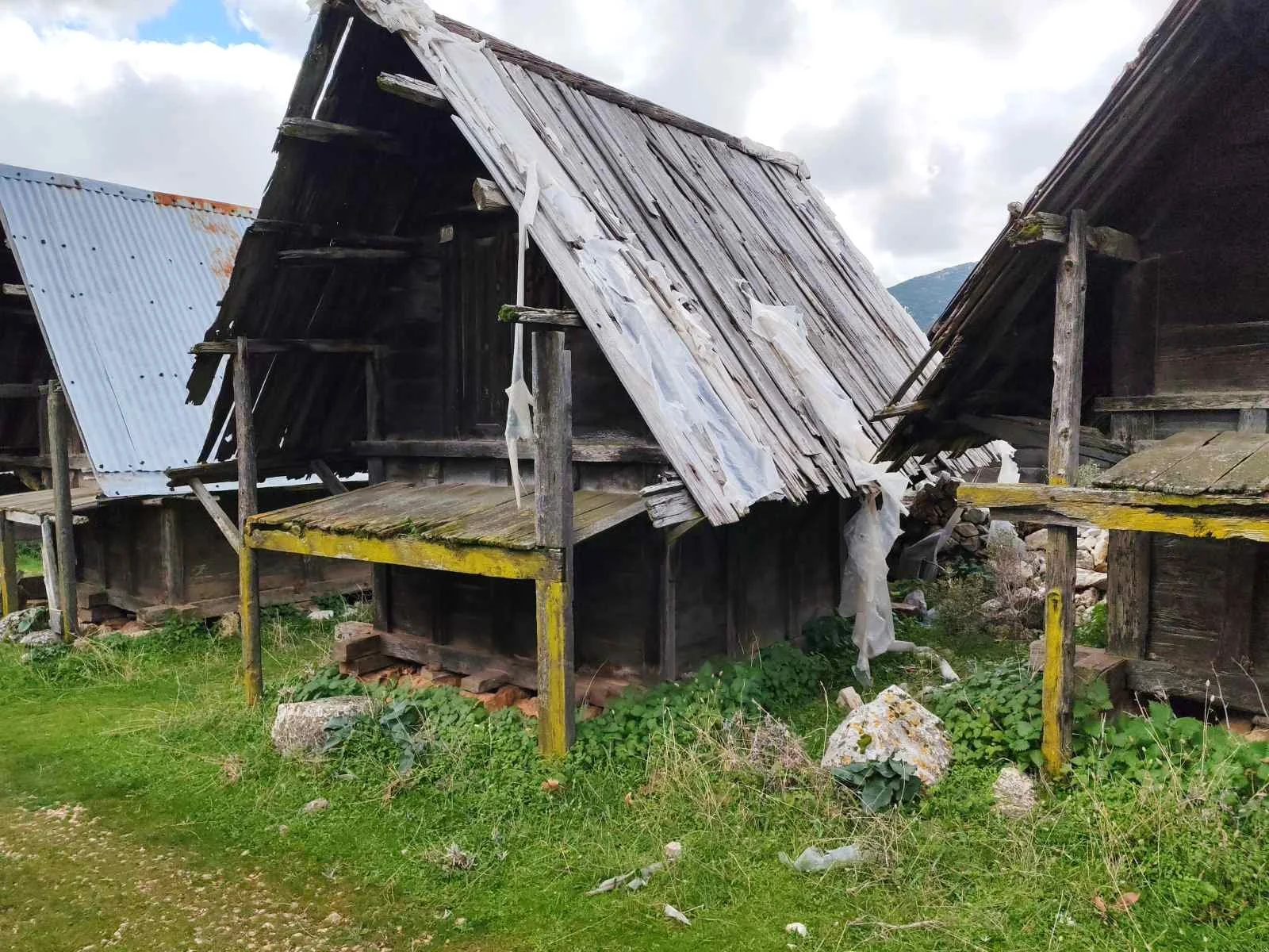 Eski ve yıkık ahşap evler, çatılarında hasar ve bozukluklar var, etrafta otlar ve taşlar bulunuyor.