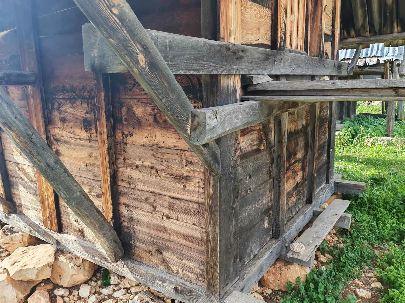 Ahşap evin düşükçe olan ahşap basamağı ve duvarı gösteriliyor, arazi üzerinde taşlar ve yeşillikler görülüyor.