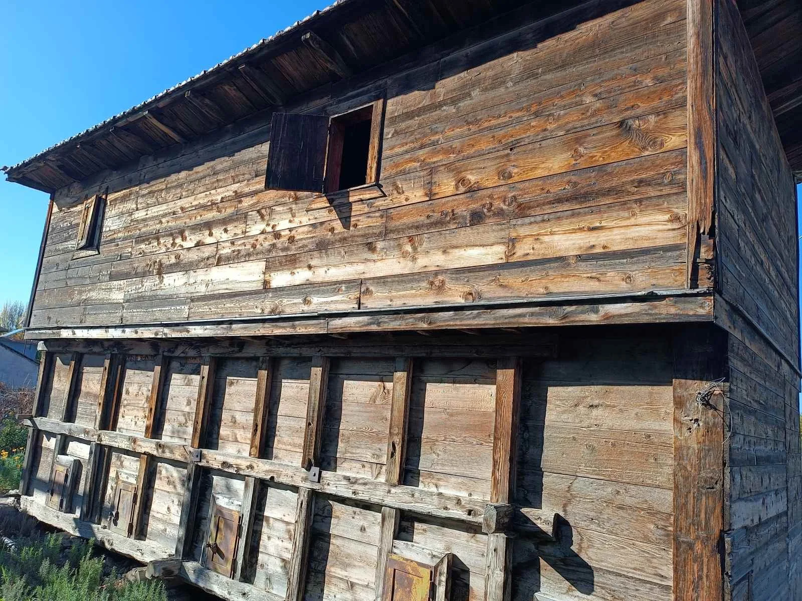 Ahşap ev, açık mavi gökyüzü altında ve bazı pencereleri açık.